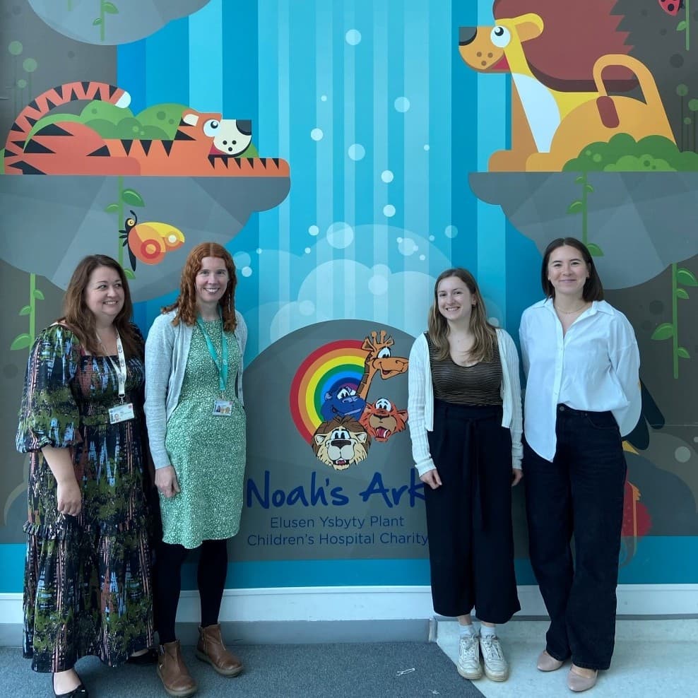 Four women standing in front of a backdrop featuring animals and a side for Noah's Ark, Children's Hospital Charity.